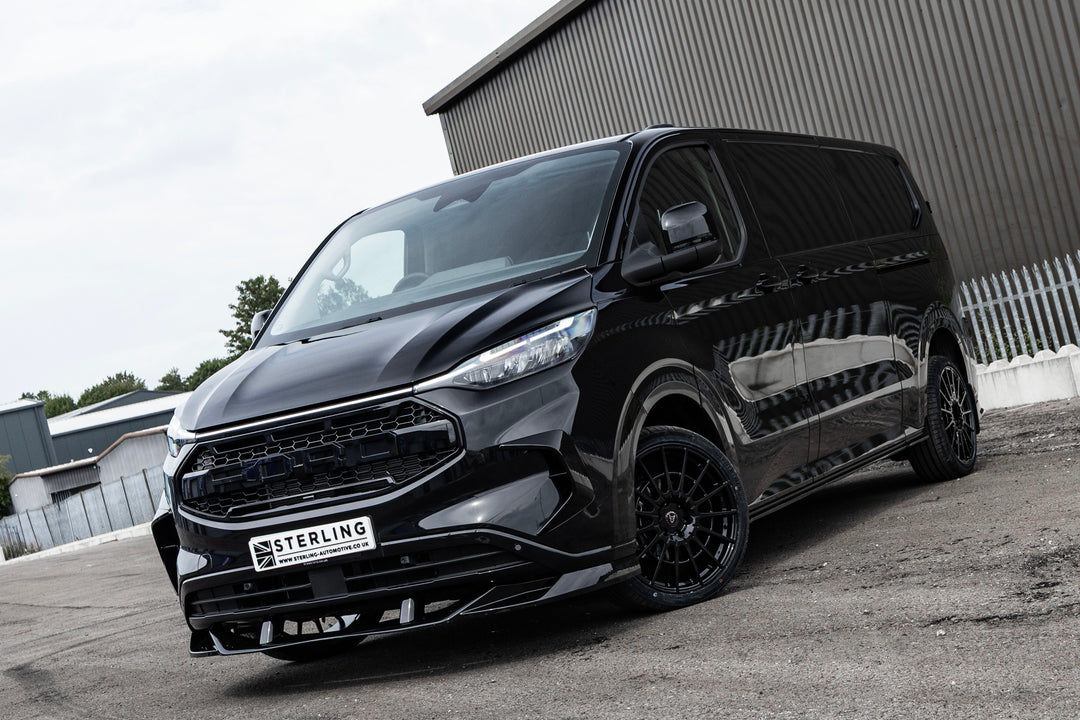 A black custom-built Ford Transit van with tinted windows and black wheels, photographed from a front three-quarter angle on an asphalt surface with a corrugated metal wall in the background.