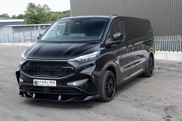 A black Ford Transit Custon FTC van with a custom front bumper and grille kit by Sterling, parked outdoors on a paved surface in front of a gray building.