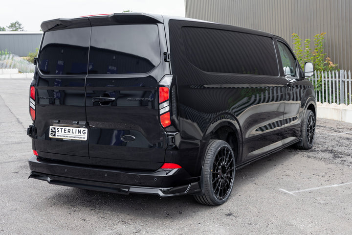 A black custom-fit Ford Transit van with black wheels, and a rear diffuser, parked on an outdoor asphalt surface.
