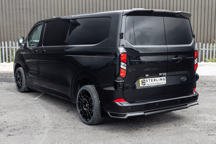 A black custom-built Ford Transit van with tinted windows and black wheels, photographed from a rear three-quarter angle on an asphalt surface with a corrugated metal wall in the background.