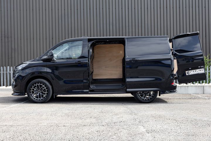 A black custom-built van with its side and rear doors open, showing a light wood interior panel, parked on an asphalt surface against a corrugated metal wall.