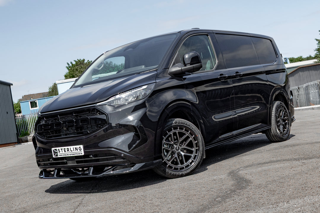 A glossy black custom-modified van with a large front grille, custom wheels, and a front lip spoiler, parked outdoors on a paved surface.