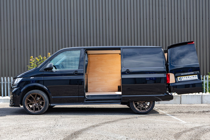 A black Volkswagen Transporter van parked outdoors with the side and rear doors open, showing a light wood interior. The vehicle is photographed in profile against a dark corrugated metal wall.