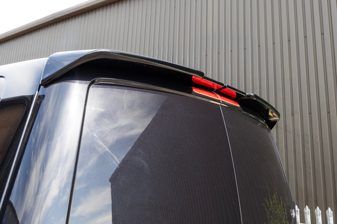 Close-up of the rear upper section of a glossy black vehicle, showing a black roof spoiler and a red rear light bar against a gray corrugated metal building background.
