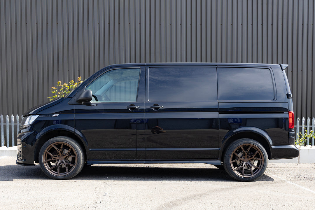 A black Volkswagen Transporter van parked in profile against a dark gray corrugated metal wall, featuring custom bronze multi-spoke wheels.