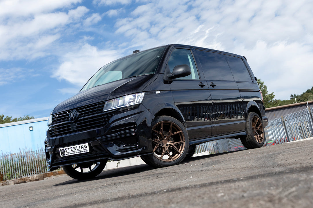 A customized black Volkswagen Transporter van with bronze-colored multi-spoke wheels, parked outdoors on a paved surface under a blue sky with clouds. The front grille features a custom black honeycomb pattern, and a license plate reading 'STERLING' is mounted on the bumper.