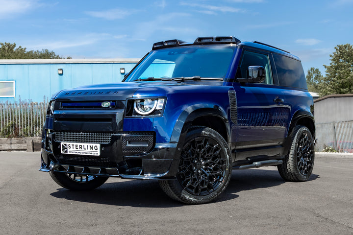 A dark blue custom-modified Land Rover Defender SUV parked outdoors on asphalt. The vehicle features a custom carbon fiber front splitter and grille by Sterling, black multi-spoke wheels, a roof-mounted light bar, and tinted glass.