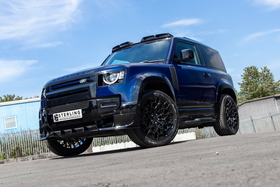 A dark blue custom Land Rover Defender SUV with black wheels and a black front grille, parked outdoors on a paved surface under a blue sky.