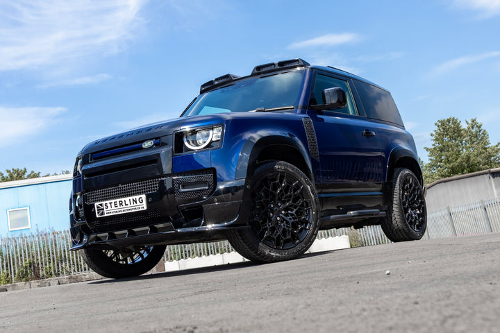 A dark blue custom Land Rover Defender SUV with black wheels and a black front grille, parked outdoors on a paved surface under a blue sky.