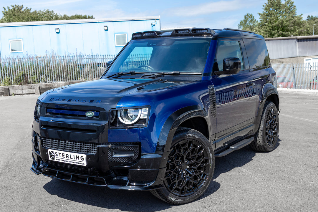 A dark metallic blue Land Rover Defender with a black custom front bumper lip kit and black multi-spoke wheels, parked on an asphalt surface in front of a light blue building.