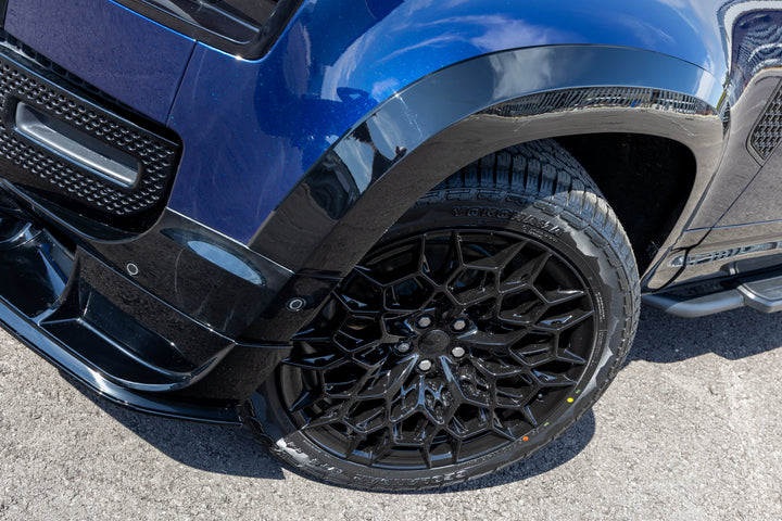 Close-up of a blue vehicle's front wheel area, showing a black multi-spoke alloy wheel and black fender overmold on a gray asphalt surface.