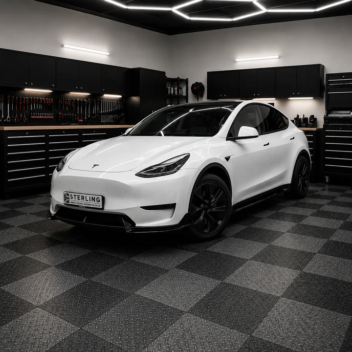 A white Tesla Model Y with black wheels and a black front lip is parked inside a modern sterling garage with a black and gray checkered floor, black tool cabinets, and hexagonal ceiling lights. A license plate reading 'STERLING' is attached to the front.