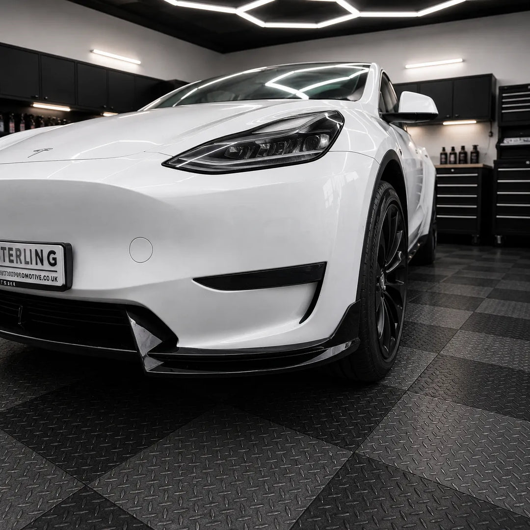 A white Tesla Model Y is shown from a low front three-quarter angle in a garage setting with a black diamond-plate checkered floor and tool cabinets in the background. The car features a custom black front lip spoiler and black wheels.