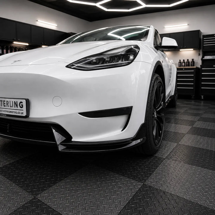 A white Tesla Model Y is shown from a low front three-quarter angle in a garage setting with a black diamond-plate checkered floor and tool cabinets in the background. The car features a custom black front lip spoiler and black wheels.
