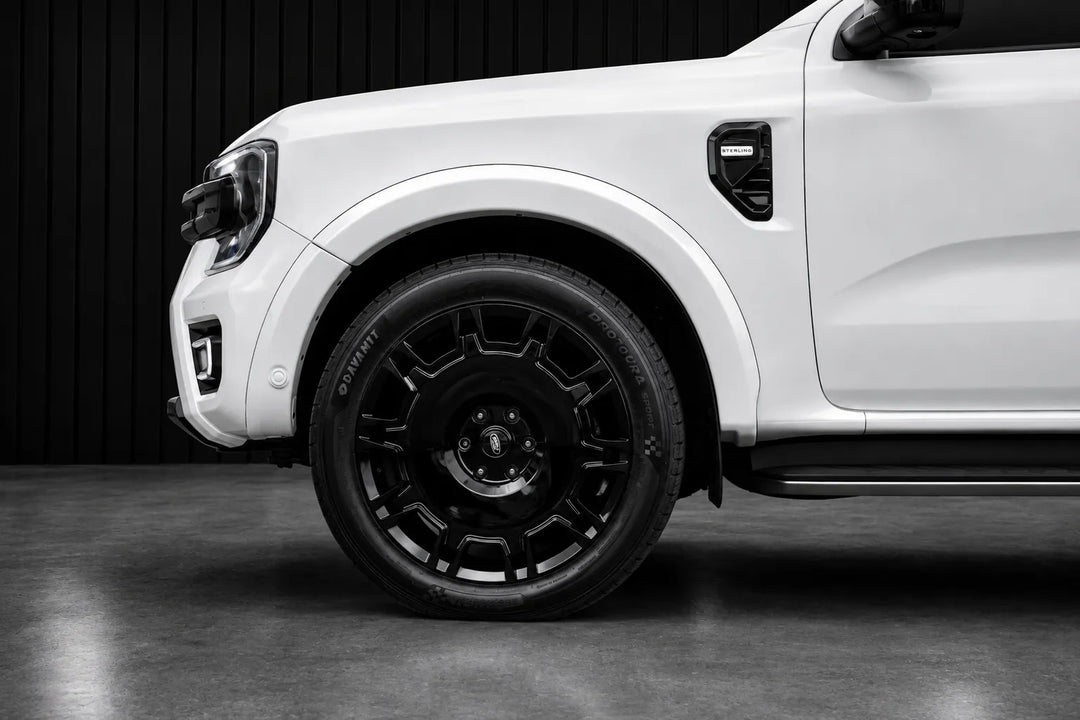 A close-up side view of a white pickup truck showing a black multi-spoke alloy wheel, black tire, and black wheel arch against a dark studio background.
