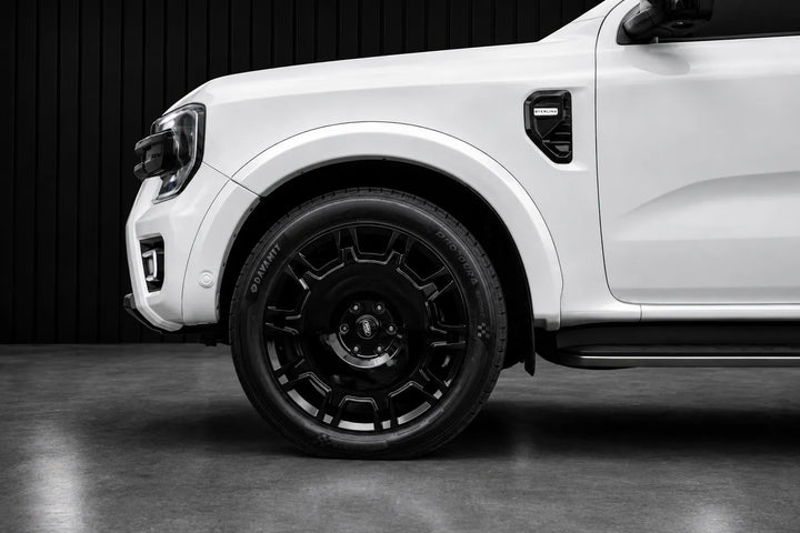 A close-up side view of a white pickup truck showing a black multi-spoke alloy wheel, black tire, and black wheel arch against a dark studio background.