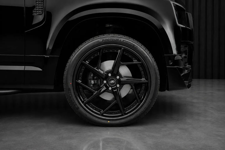 A close-up of a black multi-spoke alloy wheel with a black tire, mounted on a black vehicle, set against a dark gray studio background.