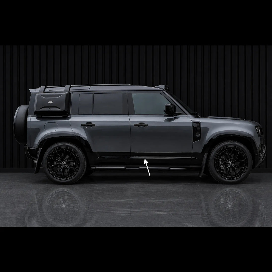 A dark gray SUV shown in a side profile view against a black slatted wall, with a white arrow pointing to the Lower Door Cladding step bars.