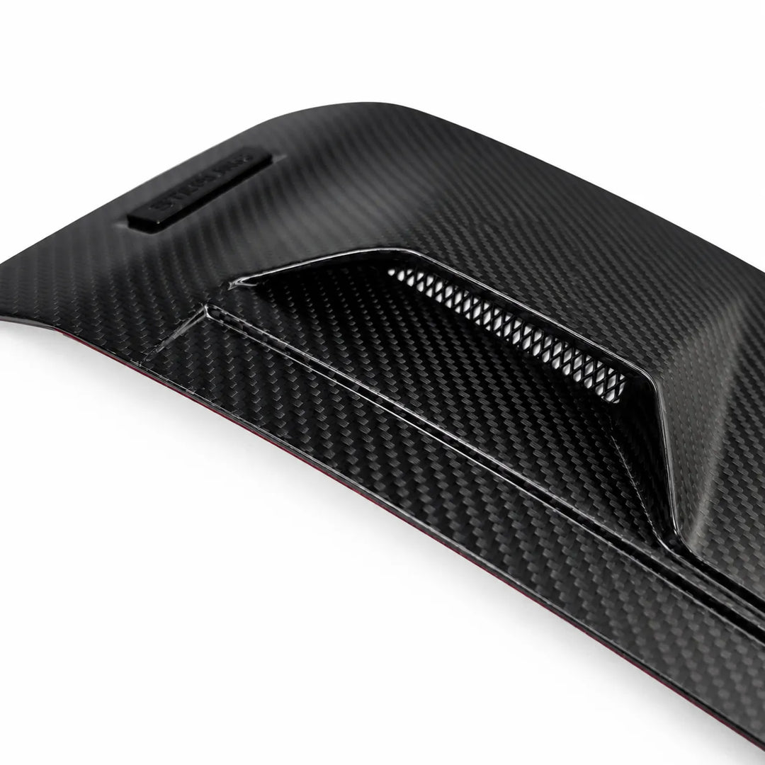 A close-up of a black dry carbon fiber textured automotive part, showing a matte woven pattern and a vented slot detail, set against a plain white background.