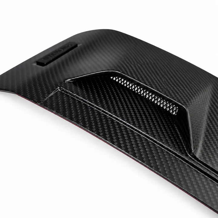 A close-up of a black dry carbon fiber textured automotive part, showing a matte woven pattern and a vented slot detail, set against a plain white background.