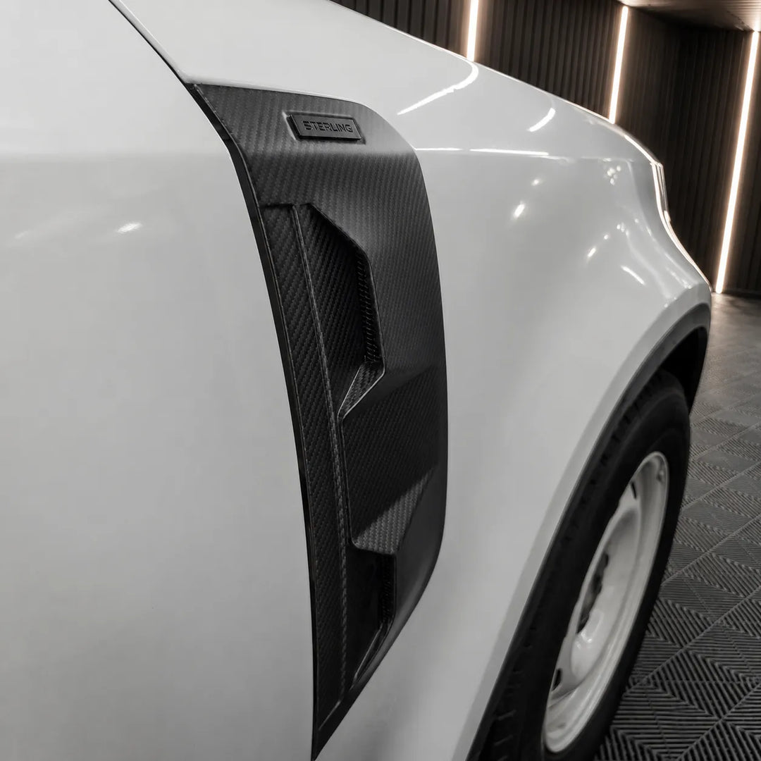 A fitted black Dry carbon fiber side Vent covers with a textured woven pattern, fitted to a land rover defender L663 displayed in a workshop background