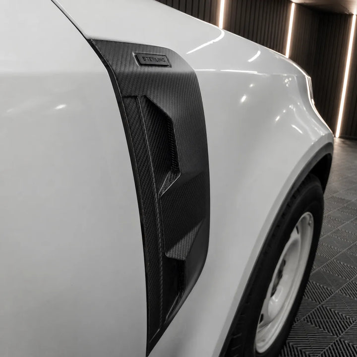 A fitted black Dry carbon fiber side Vent covers with a textured woven pattern, fitted to a land rover defender L663 displayed in a workshop background