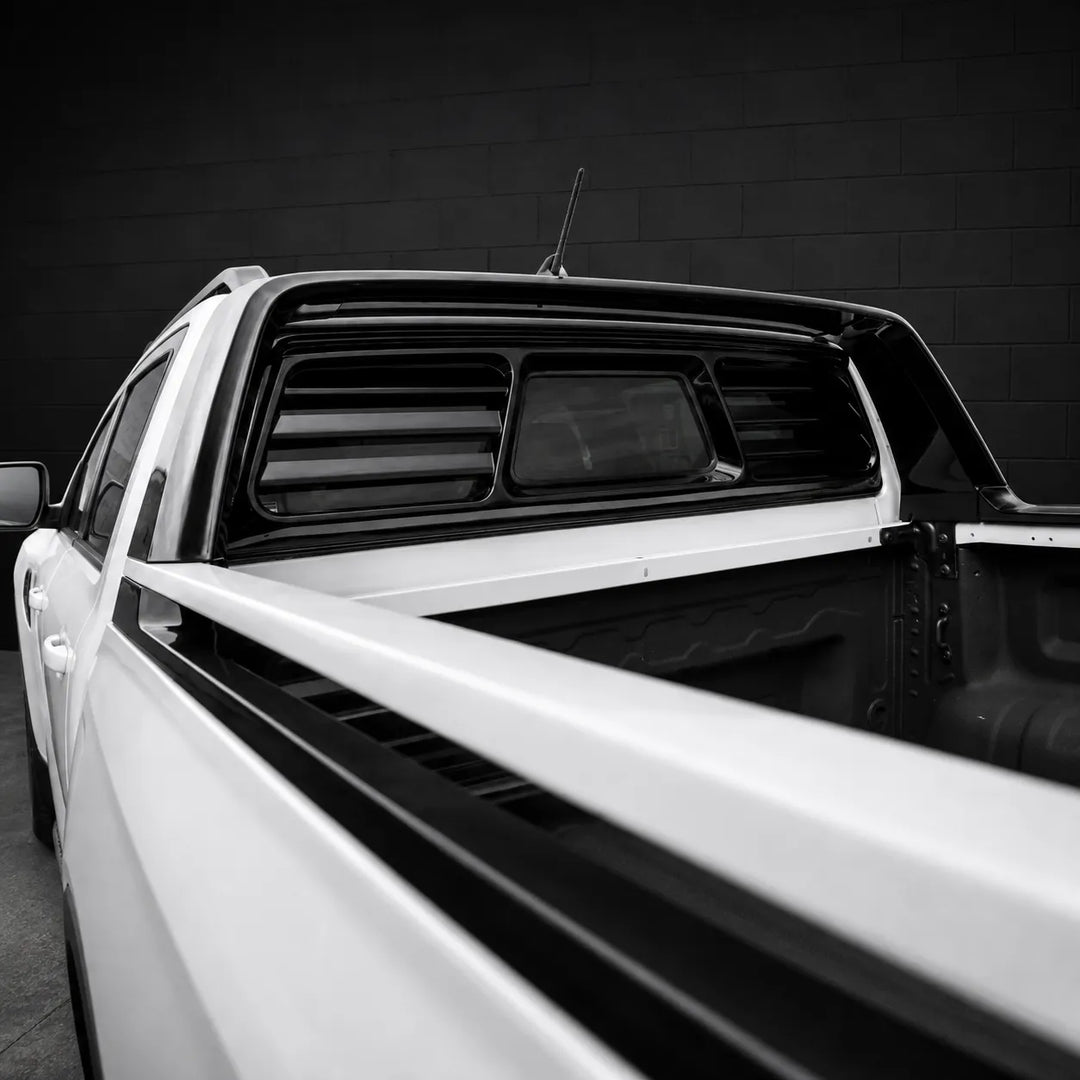 A close-up black and white photograph of the rear side of a white pickup truck bed with a black tonneau-style bed cover and rear window shutters, set against a dark brick wall.