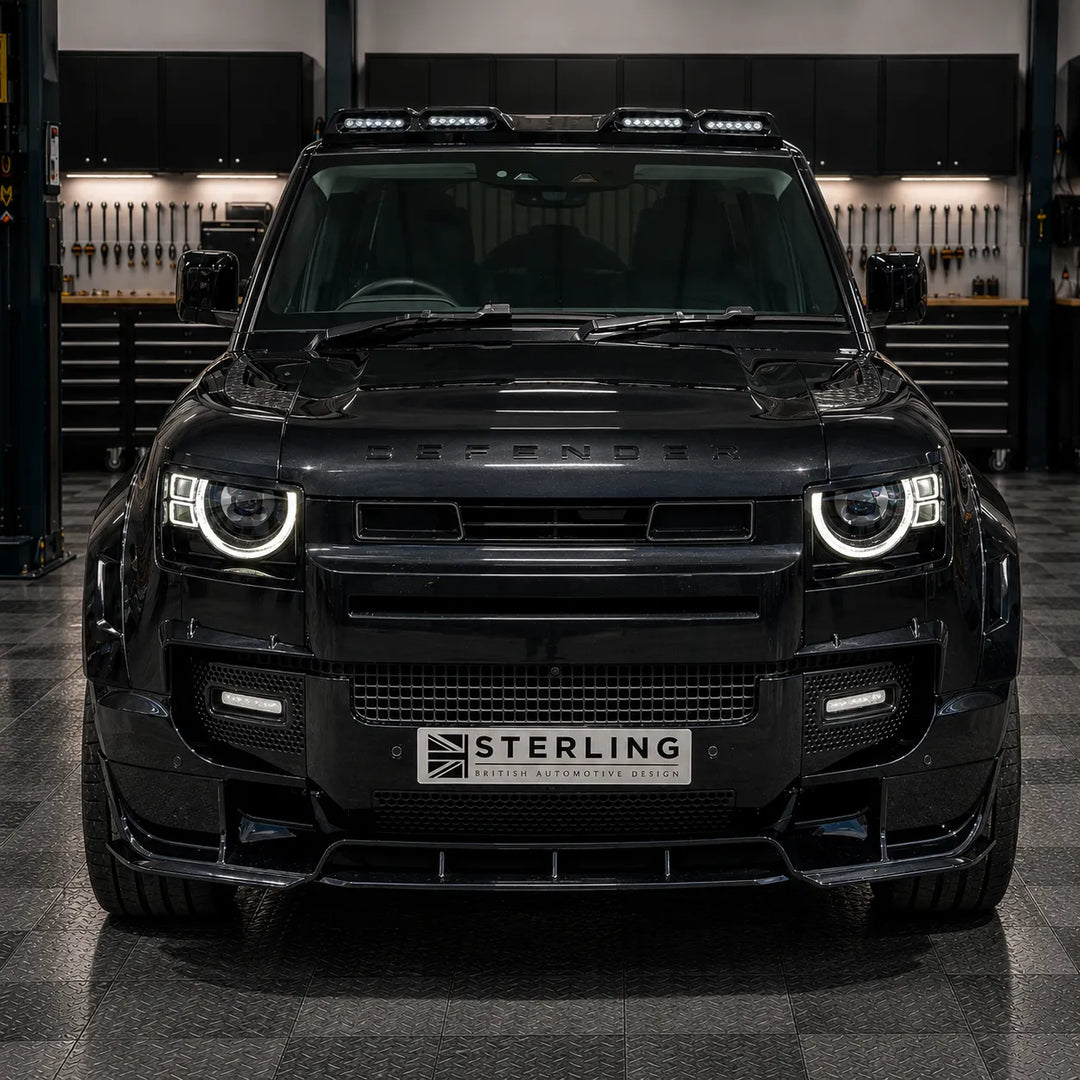 A Fitted image of the black Defender SUV parked in a workshop, featuring a custom front lip kit by Sterling with Black angular two notch front light fog surrounds winglets. The vehicle has a black grille surround, integrated fog lights, and a license plate frame with the 'STERLING' logo.