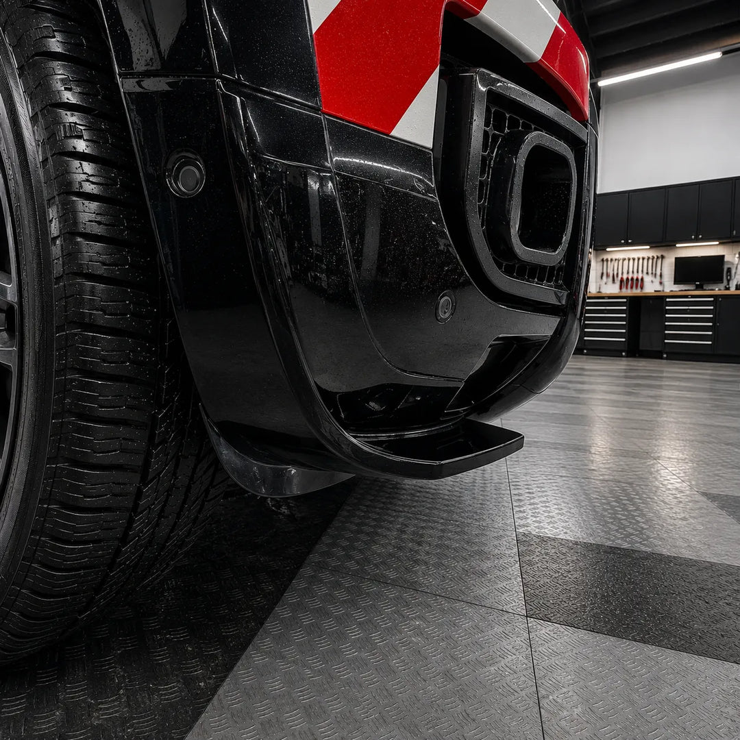 Close-up of a black vehicle front bumper fitted glossy black lower automotive front bumper lip trim canard spoilers, The image is photographed in a garage with a diamond-plate floor.