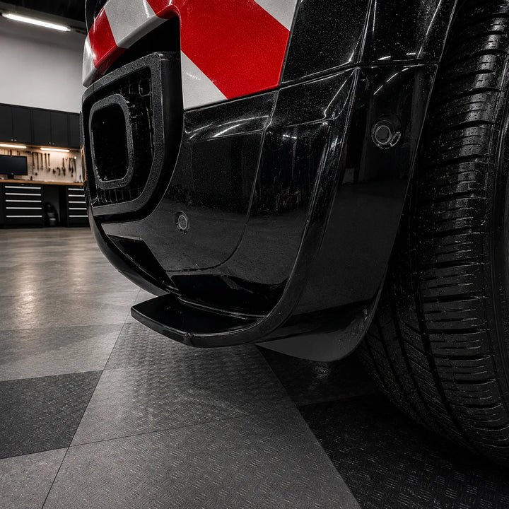 Close-up of a glossy black defender front bumper lower area fitted with aggressive canards in a glossy finish, with a wet diamond-plate floor and a workshop visible in the background.