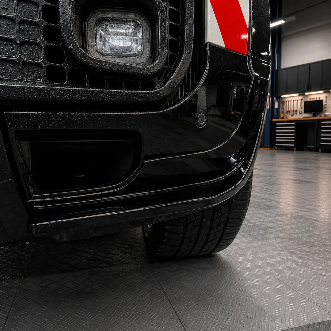 Close-up of a glossy black defender front bumper lower area fitted with aggressive canards in a glossy finish, with a wet diamond-plate floor and a workshop visible in the background.