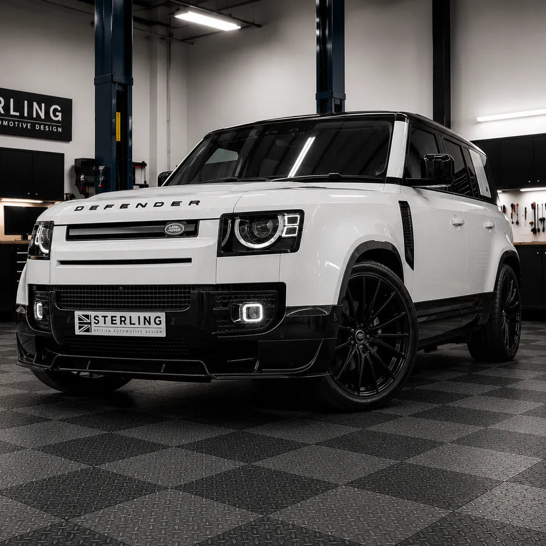 A white Land Rover Defender SUV with custom Sterling Design black wheels and a black front lip kit, parked inside a the sterling workshop with a checkered floor and a Sterling British Automotive Design sign on the wall.