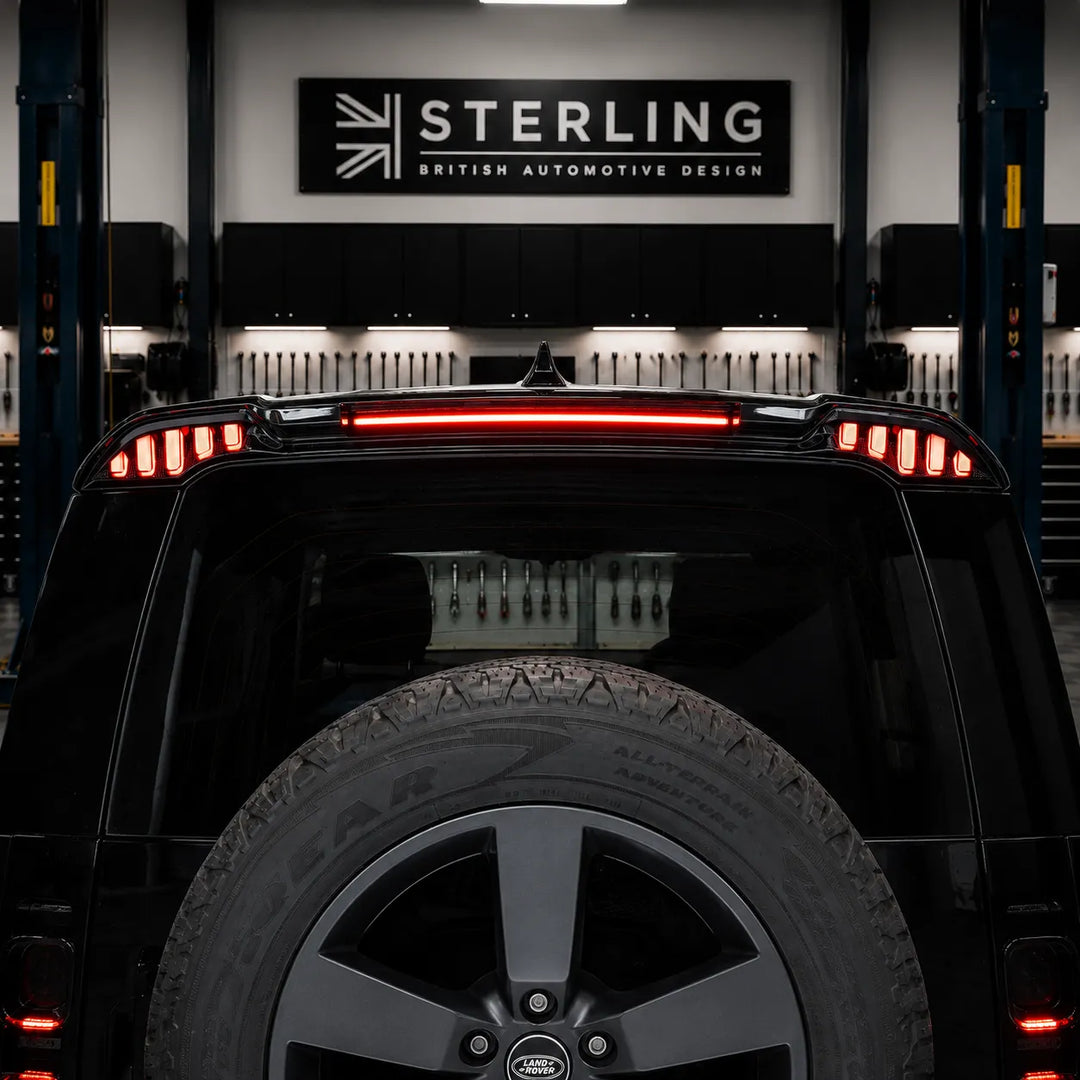 A black automotive rear spoiler with integrated LED light bars, shown both as a standalone product on a white background and installed on the rear of a black land rover defender with led lights spoiler lip shot in the sterling garage setting.