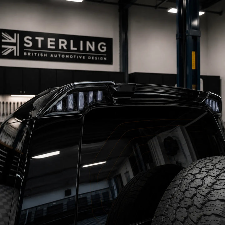 Close-up of a glossy black defender l663 led spoiler rear section in sterling garage, with a large tire visible in the lower right and a Sterling British Automotive Design sign on the wall behind.