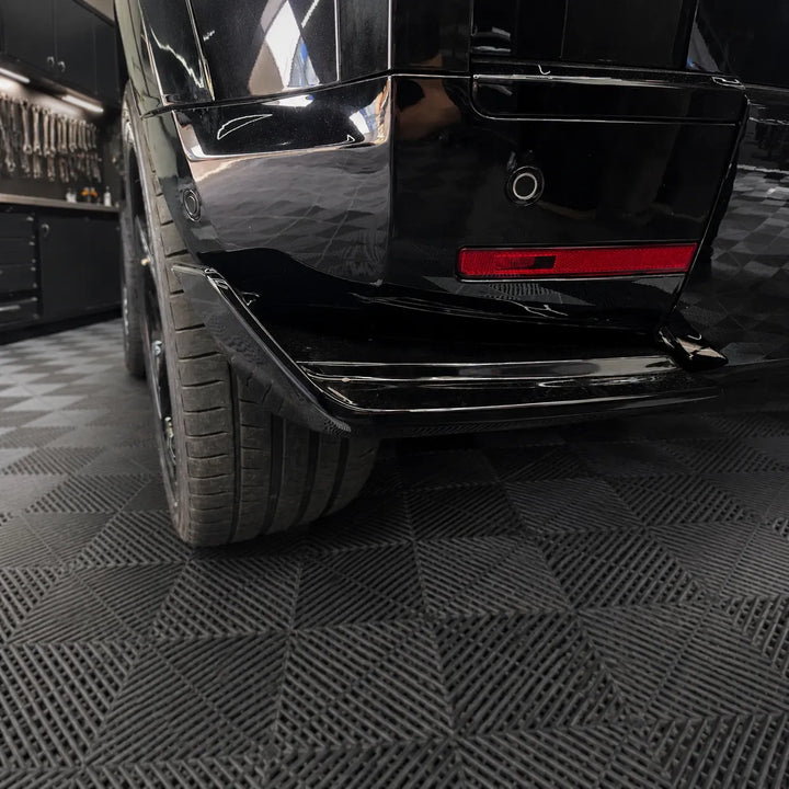 A close-up of a glossy black vehicle rear bumper and lower bumper area with low canards installed on a black Defender L663, photographed the sterling garage with dark textured floor tiles.