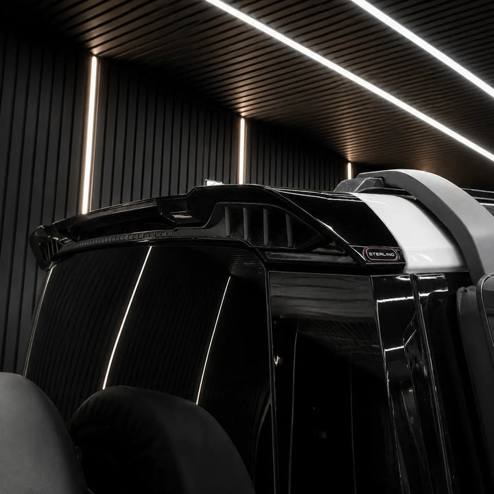 Close-up of a glossy black vehicle rear section with a small visible Sterling badge, photographed in a dark modern interior with linear ceiling lights.