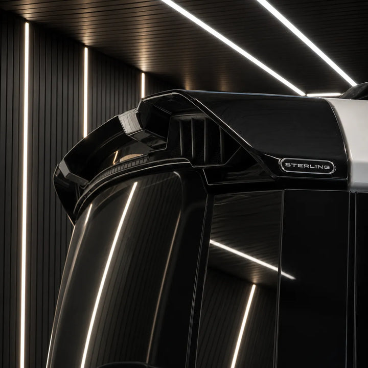 Close-up of a glossy black vehicle body panel with a visible 'STERLING' badge, photographed in a modern interior with vertical LED strip lighting.