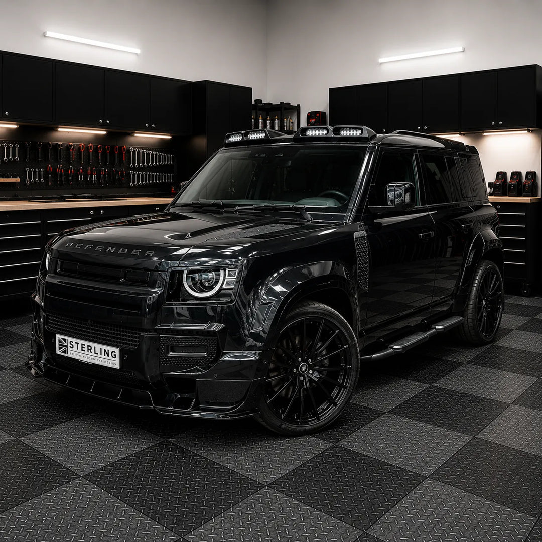 A black custom-modified Defender SUV parked inside the sterling workshop garage with a black and gray checkered floor. The vehicle features a custom front lip, a license plate reading 'STERLING', and black multi-spoke wheels.