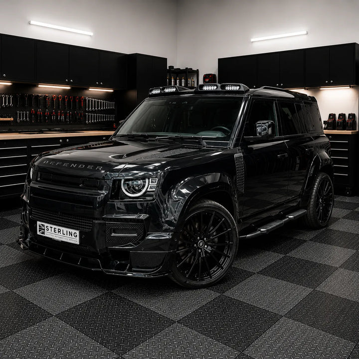 A black custom-modified Defender SUV parked inside the sterling workshop garage with a black and gray checkered floor. The vehicle features a custom front lip, a license plate reading 'STERLING', and black multi-spoke wheels.