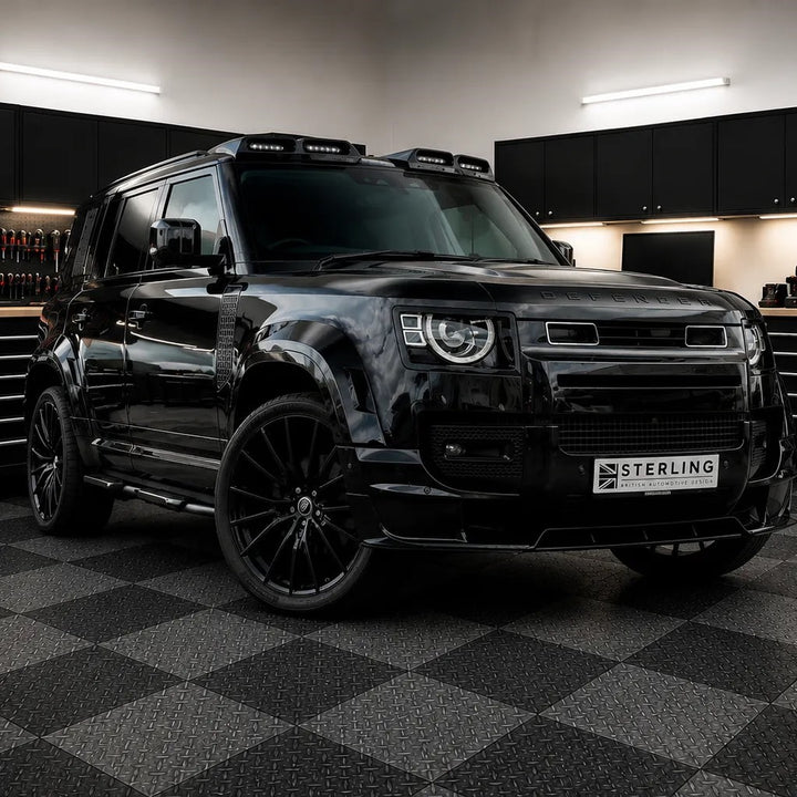 A glossy black Land Ropver Defender L663 parked in teh sterling workshop on a black-and-gray checkered floor, with a visible Sterling badge on the front license plate area and a dark cabinetry wall in the background. Fitted is the Wide Wheel Arches finished in gloss black

