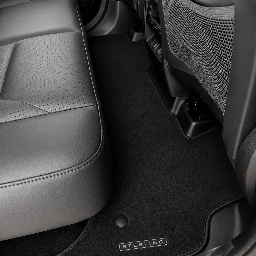 Black car floor mats installed inside a vehicle, shown around the front seat and between the front and rear seats. A small metal badge on the mat reads STERLING.