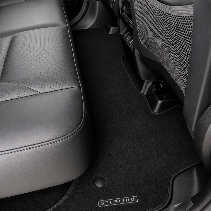 Black car floor mats installed inside a vehicle, shown around the front seat and between the front and rear seats. A small metal badge on the mat reads STERLING.
