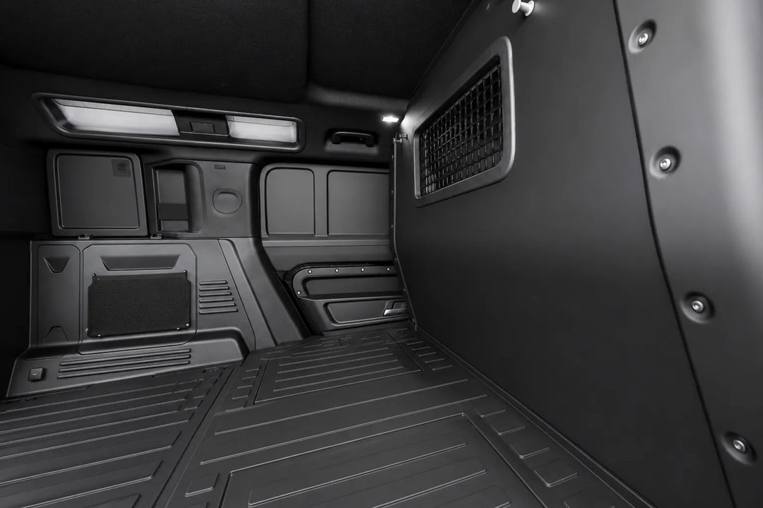 The interior cargo area of a black Defender van, showing a textured floor mat, side wall panels, and a storage compartment on the left.