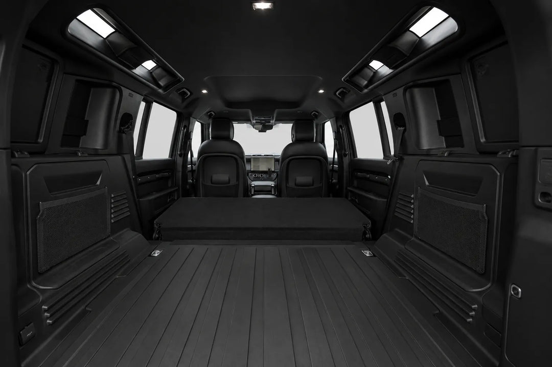 The interior cargo area of a black van with a fold-down rear bench, black floor mats, and side cargo pockets, shown from a rear-facing perspective.