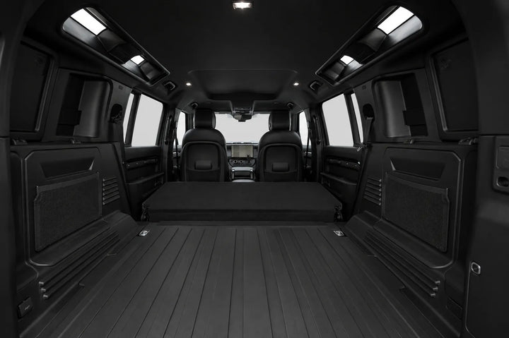 The interior cargo area of a black van with a fold-down rear bench, black floor mats, and side cargo pockets, shown from a rear-facing perspective.