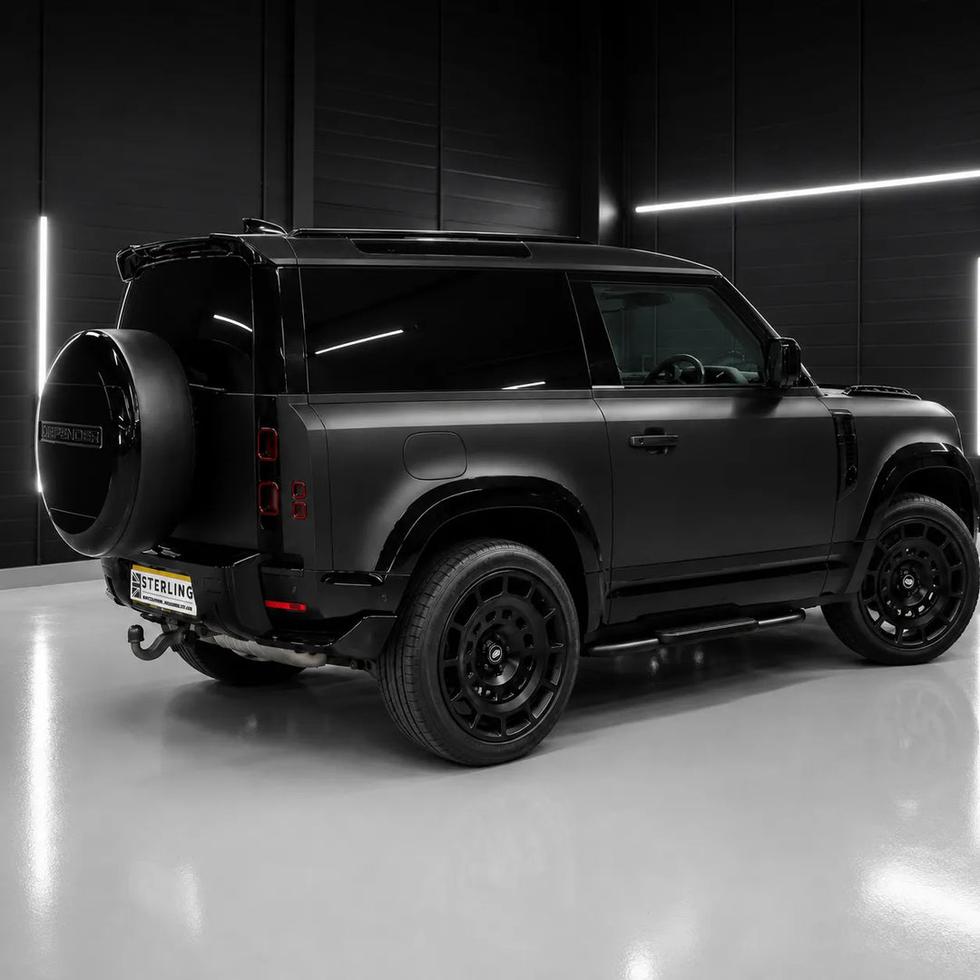 A matte black SUV shown from a rear three-quarter angle in a dark studio setting with bright linear lighting on a reflective gray floor.