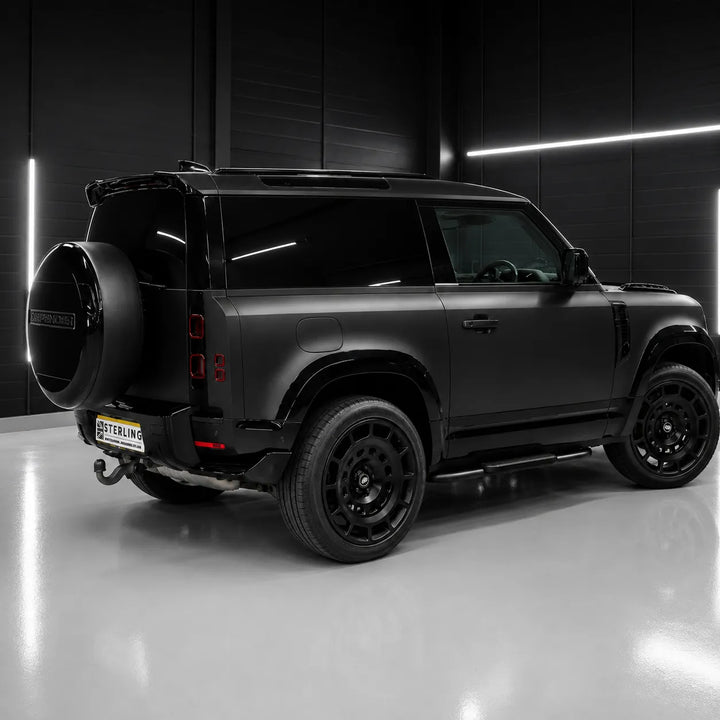 A matte black SUV shown from a rear three-quarter angle in a dark studio setting with bright linear lighting on a reflective gray floor.