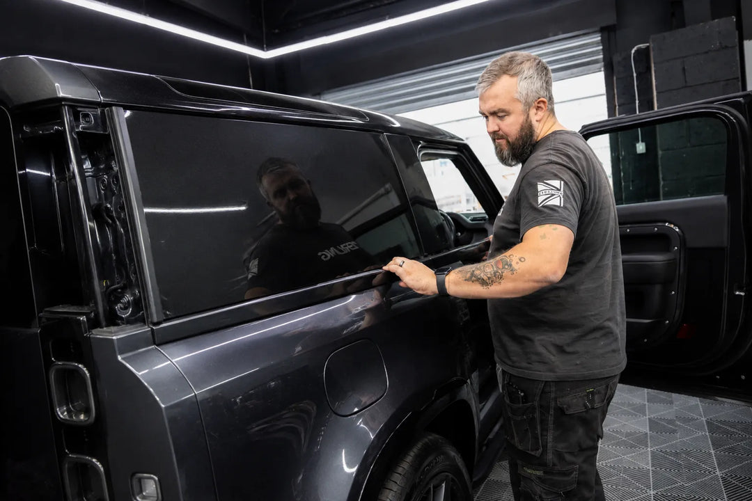 A man with a beard and tattoos stands next to a dark gray SUV in a garage, touching the window trim. The vehicle has tinted glass and a glossy finish, with its side door open.