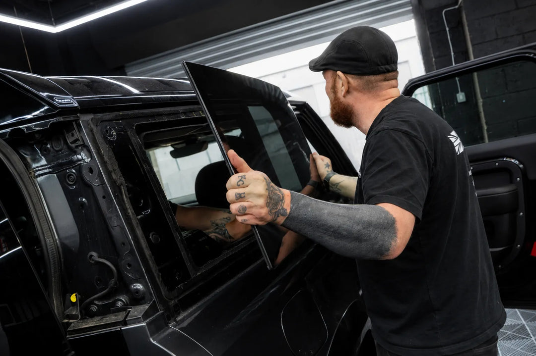 A tattooed man in a black cap and black t-shirt installs a tinted glass window panel onto a black Land Rover Defender 90 inside the sterling workshop.