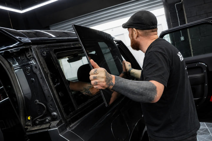 A tattooed man in a black cap and black t-shirt installs a tinted glass window panel onto a black Land Rover Defender 90 inside the sterling workshop.